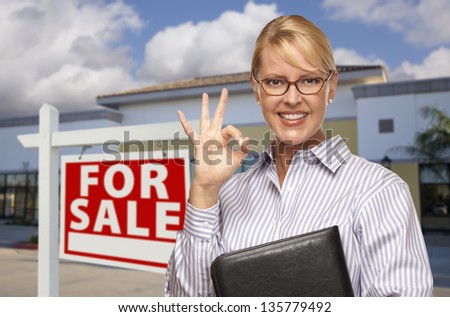 Smiling Businesswoman with Okay Sign In Front of Vacant Office Building and For Sale Real Estate Sign.