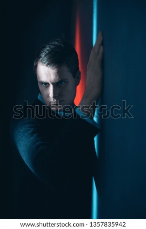 Suspicious man in black sweater stands against wall in red and blue light.