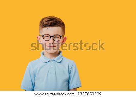 portrait of a beautiful and handsome little boy who looks into the camera and smiles, in glasses, dressed in blue shirt, isolated on yellow background, copy space Royalty-Free Stock Photo #1357859309