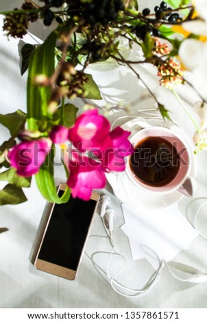 
Concept of female good morning. Pink cup of tea or coffee, on a plate of candy, a smartphone, headphones, sheets for writing, a bouquet of flowers. Light background. Selective focus.