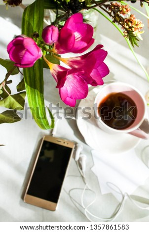 
Concept of female good morning. Pink cup of tea or coffee, on a plate of candy, a smartphone, headphones, sheets for writing, a bouquet of flowers. Light background. Selective focus.
