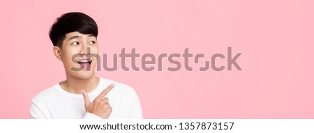 Smiling young handnsome Asian man pointing hand to empty space aside on pink banner background