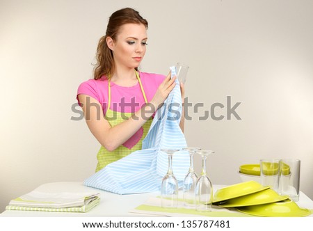 Beautiful young woman wipes clean utensils in kitchen on grey background