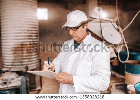 Serious Caucasian veterinarian in white uniform writing down results of animal examination while standing in front of barn.