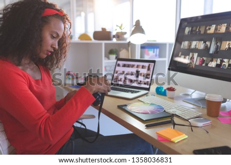 Side view of young mixed-race female graphic designer reviewing photos in digital camera at desk in a modern office