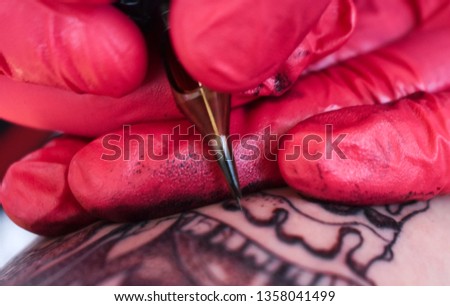 Close up macro of hands tattooing an arm wearing red medical gloves and using black ink in a tattoo studio.