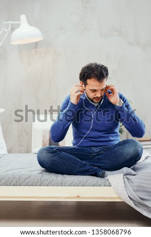 Attractive arab man sitting on bed in bedroom dressed casual and putting earphones in ears.