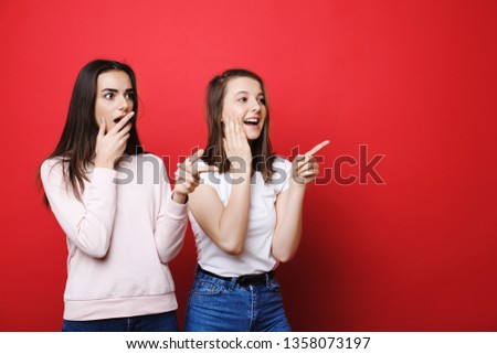 Two lovely young caucasian woman looking away amazed and pointing away against red background.