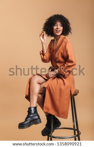 Vertical image of Cheerful pretty african woman sitting on chair and looking away over brown background