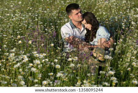 Happy and carefree lovers in nature. Emotional portrait of a beautiful and pensive girl hugging with her boyfriend sitting in a field of daisies at sunset. Summertime. Summer vacation. Lifestyle