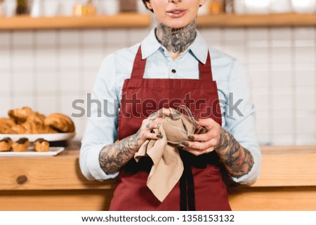 partial view of barista in apron wiping glass cup with rag  