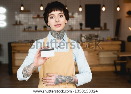 attractive waitress in apron holding credit card and looking at camera