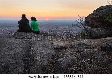 Young romantic couple sitting on mountain cliff looking to stunning panorama over small town Vrsac in sunset.