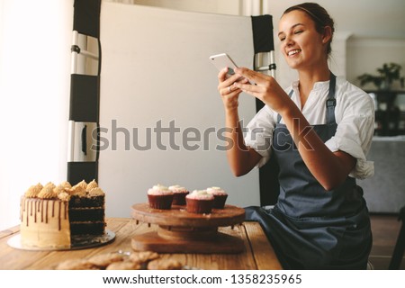 Happy woman chef taking a picture of pastries on a wooden board with smart phone while standing at the kitchen. Female baker taking pictures of the dessert for her food blog.