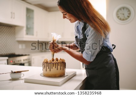 Female chef decorating cake with whipped cream using party bag. Woman in apron preparing a delicious cake at home.