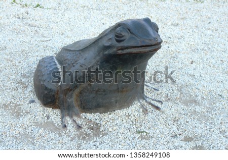 A large stone frog sits on white stones in the park. Sculpture of black toad made of metal.