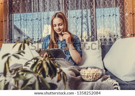 Girl sitting on the sofa covered with a blanket at home with popcorn, watching movie on tablet. - Image