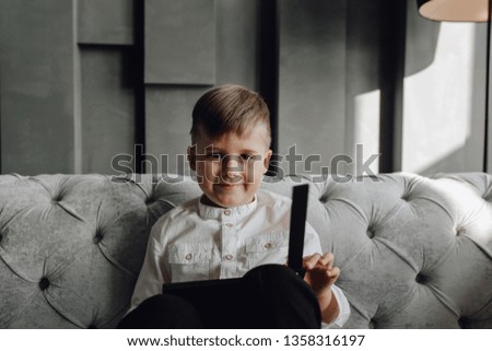 Little Boy with Clapper Board on Couch Medium Shot. Caucasian Blond Kid in White Shirt Sit on Grey Sofa, Smile Head and Shoulder Photo. Child with Brown Eye Looking at Camera Front View