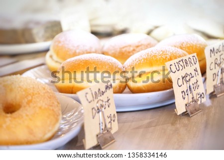 bright, soft shot of variety favors of donuts, each type put separately on white dish on wooden table. In front are price tags for each. Selective focus on custard cream donut. 
