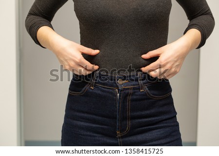 A closeup view of a young Caucasian woman pinching the thin layer of fat around her waistline, just above her well-fitted denim jeans. Body dysmorphia concept. Royalty-Free Stock Photo #1358415725