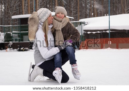 Smiling young mother and her cute daughter ice skating together. Family skating and training with white skates on the ice area in winter day. Weekends activities outdoor in cold weather