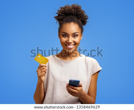 Smiling girl holds smart phone and credit card. Photo of african american girl wears casual outfit on blue background. Emotions and pleasant feelings concept.