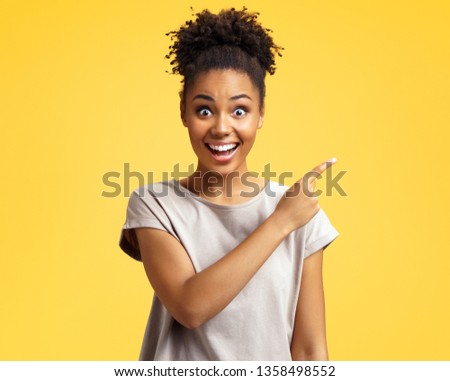 Overjoyed girl points aside, shows something at freespace. Photo of african american girl wears casual outfit on yellow background. Emotions and pleasant feelings concept.