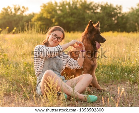 Pretty brunette female girl making hands in heart shape with a dog German Shepherd on the grass in park. Young woman with her dog on green meadow