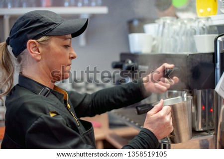 Middle Aged Woman Dressed in Black Kitchen Uniform Wearing a Baseball Cap Making Coffee using a Barista Machine with Steam Arising from a Chrome Jug for Heating up the Milk.