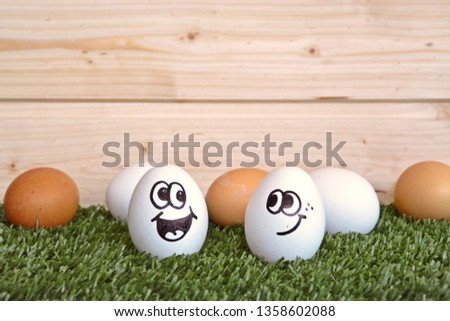 Two white eggs with funny faces drawn on it lie on a lawn with a higher background and other brown and white eggs behind it