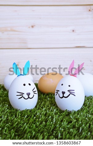 Two eggs painted with the face of a hare and glued to rabbit ears lie on a grass field in front of a wooden background with space for text or other elements