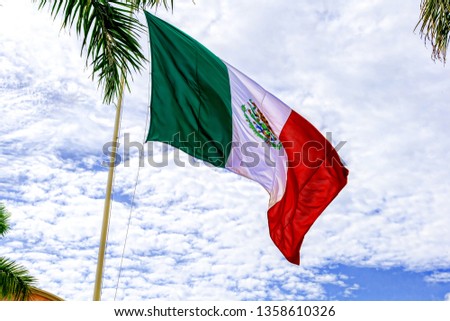 The flag of Mexico develops in the wind against a blue sky and tropical trees. United States of Mexico.