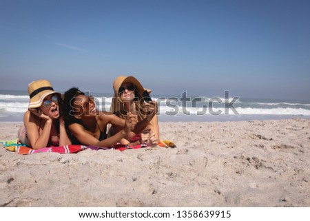 Front view of happy beautiful young multi ethnic women with hat and sunglasses taking a selfie with a mobile phone while lying on a blanket on the beach 