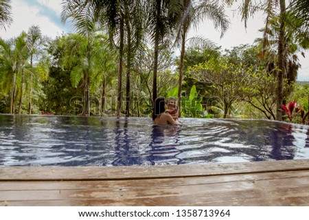 A young mother with her beautiful daughter, with curly hair, in an infinity pool, in the middle of nature, in the recreation area of a resort, outdoors and with native plant garden.