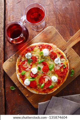 Overhead view of a colourful tricolore Italian pizza with cheese, tomatoes and fresh basil on a wooden cutting board served with a jug and glass of red wine on an old table. More pizza at my port.