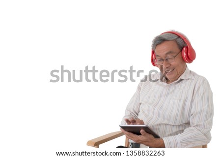 An Asian old man sitting on a chair and enjoying using a tablet with red headphone listening to music on isolated white background