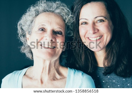 Smiling senior woman and her daughter posing at camera. Mother and daughter with grey wall in background. Family portrait concept. Front view.