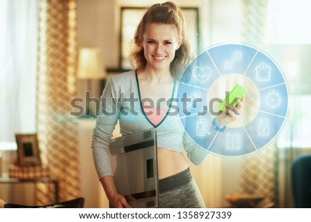 Portrait of smiling fit woman in fitness clothes with smart scale tracking weight in a fitness app in a smartphone at modern home.