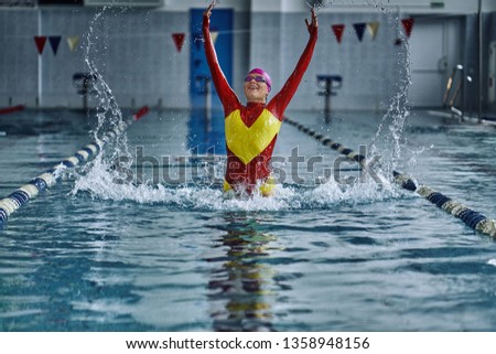 Female athlete in a red-yellow swimsuit makes a jump for joy in the pool. Splashes of water scatter in different directions.