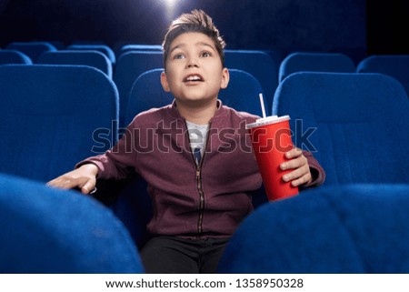 Shocked, fascinated boy sitting in movie theatre, looking at screen attentively with opened mouth. Teenager holding red paper cup with fizzy drink.