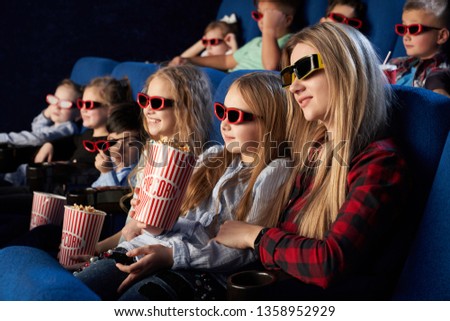 Mother and daughter sitting together in movie theatre, enjoying premiere of film. Children wearing in 3D glasses watching movie, holding popcorn buckets.