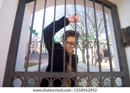 The young man with a smile poses, standing behind the iron bars of the vintage gate. Dressed in a black light overcoat, black-rimmed glasses.