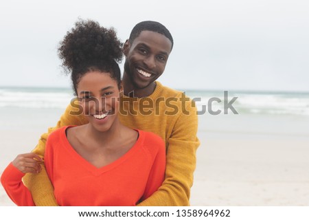Portrait of diverse couple standing at beach on a sunny day. They are smiling and looking at camera Royalty-Free Stock Photo #1358964962