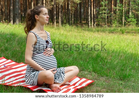Portrait of a beautiful pregnant girl meditating with closed eyes sitting on a rug on the grass in a forest clearing on the background of trees and hugging a big tummy.