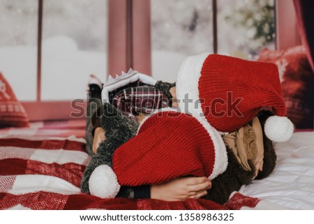 Two little brothers in red christmas hats playing hugging wrestling on bed with red tartan blanket, blurred window on background.