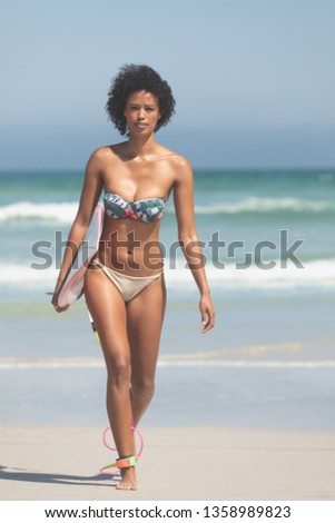Portrait of beautiful Mixed-race female surfer with a surfboard walking at beach on a sunny day. She is looking at camera