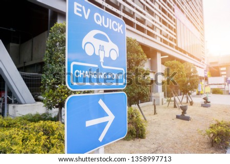 Perspective view and closeup sign and symbol of Electric car charging point with navigation arrow on blurry building and sun flare background.