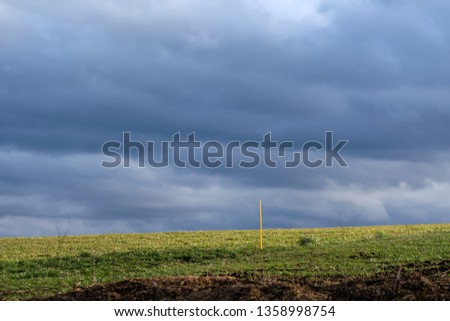 Landscape in countryside with grey overcast sky but also sunlight illuminating the green grass on the fields - and some yellow poles sticking in the ground. Seen near Heroldsberg, Germany, March 2019
