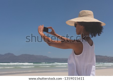 Side view of pretty young mixed-race woman talking selfie at beach on a sunny day. She wears hat and sunglasses