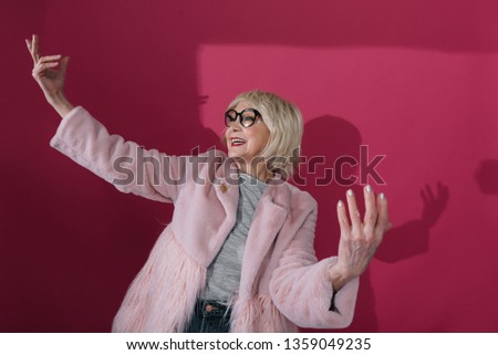 Cropped photo of happy stylish old lady in fashionable pink jacket with raising hands up against red wall. She is looking aside while posing for camera indoors
